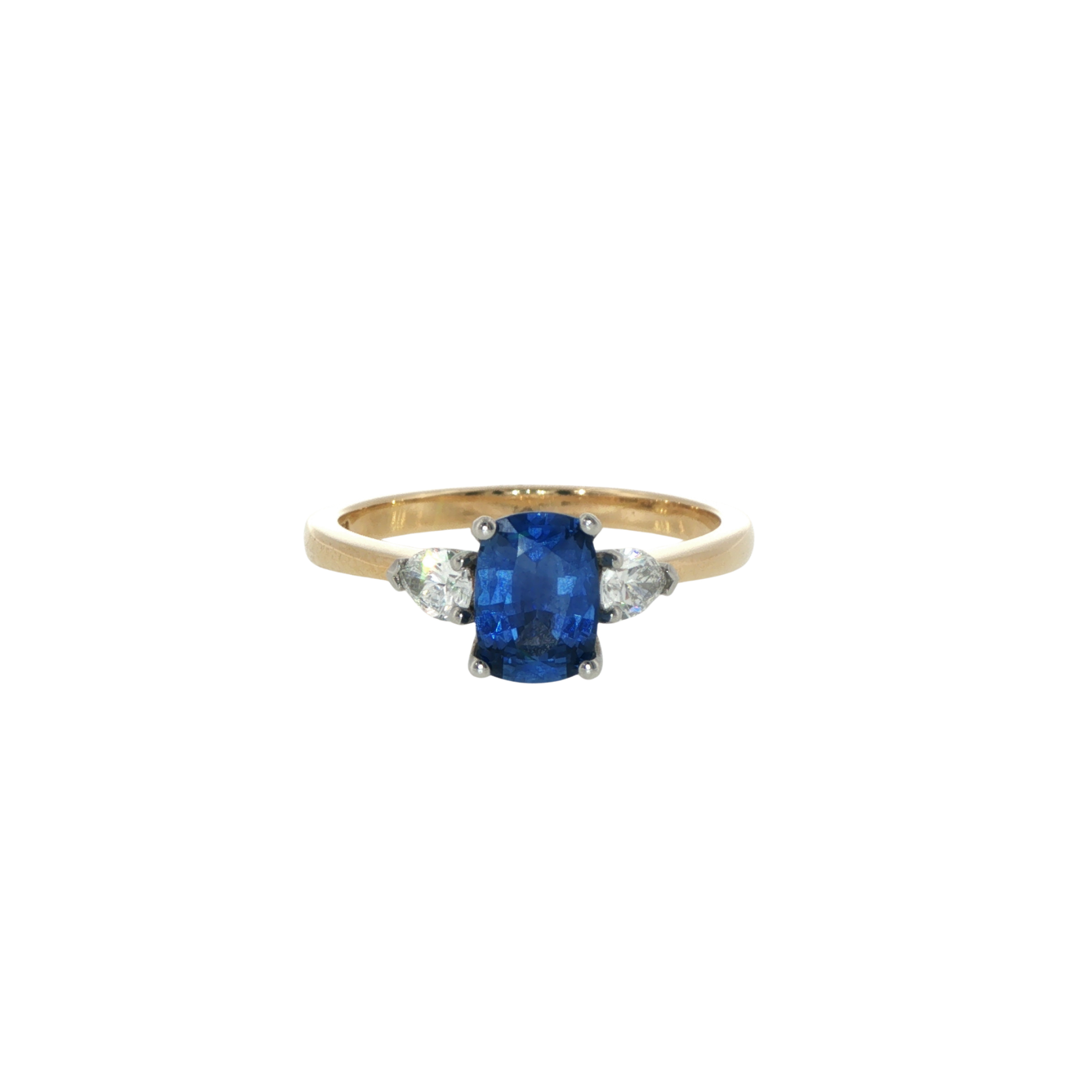 A sapphire trilogy engagement ring. The centre sapphire is blue and cushion cut, flanked by two pear cut diamonds. They are on a yellow gold band and set with platinum claws. The ring is lying flat, face on, on a white background.